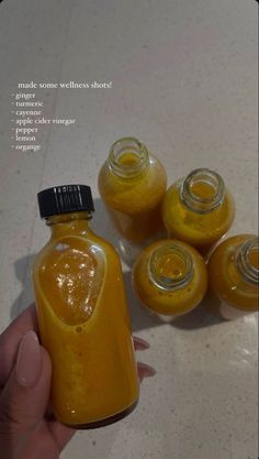 a hand holding a jar filled with yellow liquid next to five small jars full of orange juice