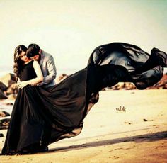 a man and woman kissing on the beach while wearing long black dresses with flowing fabric