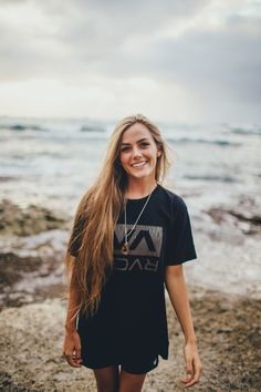 a woman standing on top of a rocky beach next to the ocean with her long hair blowing in the wind