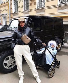 a woman standing next to a baby in a stroller near a black truck and buildings