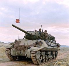 two men riding on top of a tank in the middle of a dirt road with an orange flag