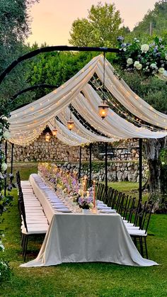 an outdoor dining table set up with white linens and lights on the top, surrounded by greenery