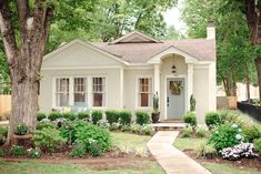 a small white house with trees and flowers in the front yard on a sunny day