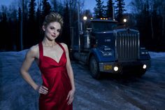 a woman in a red dress standing next to a semi truck on a snowy road