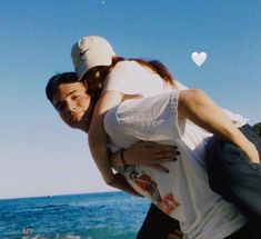 a man holding a woman on his back near the ocean with a heart shaped object in the background