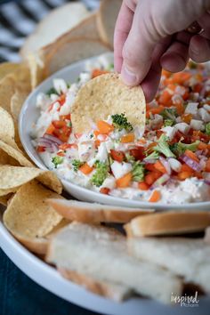 a hand dipping a tortilla chip into a bowl of dip with veggies and cheese