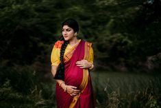 a woman in a red and yellow sari standing next to some tall grass with trees in the background