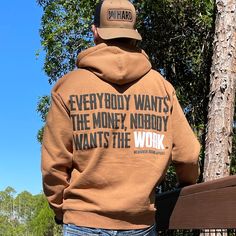 Man pictured from back wearing a WW Saddle Brown Hoodie. EVERYBODY WANT$ THE MONEY, NOBODY WANTS THE WORK printed in bold on full back width in black ink, except the word WORK is printed in white for emphasis. (WEWORKIN BRAND APPAREL printed small and lower right just below the full back imprint.) Banded cuffs and waist. Hood down on back. (Also wearing a WW HARD Coyote Brown/Black Retro Trucker hat backwards.) Brown Hoodie, Saddle Brown, Hoodies Mens, Lifestyle Clothing, Get The Job, Mens Graphic Tee, The Money, In The Woods, Heavy Weight