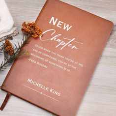 a brown book sitting on top of a wooden table next to flowers and a white towel