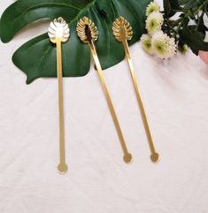 three gold hair pins sitting on top of a white table next to flowers and leaves