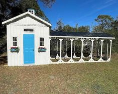 a small white building with blue door and windows on the outside, in front of some trees