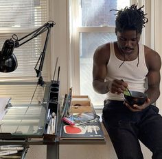 a man with dreadlocks sitting at a desk in front of a window looking at his cell phone