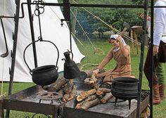 a woman cooking food over an open fire in a field with other people standing around