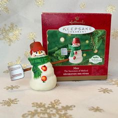 a small snowman with a red hat and green scarf next to a boxed keepsake christmas ornament