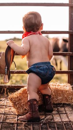 a toddler in cowboy boots and bandana sitting on hay next to a fence