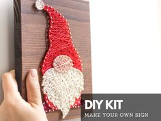 a hand is holding the string art santa clause on a wooden plaque with red and white yarn