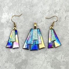 three pieces of glass are hanging from gold earwires on a gray surface, one is multicolored and the other is blue