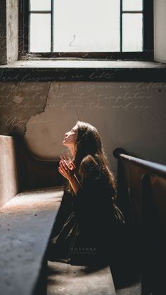 a woman sitting in front of a window with her hands clasped to her chest and looking up