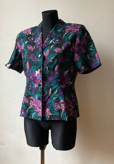 "♥ Vintage short sleeve blouse, button down. ♥ Floral pattern in purple, violet, green and black. ♥ Beautiful collar, deep v neck, padded shoulders. ♥ 100 % Thai silk.  ♥ Good vintage condition. ♥ Brand: Shinawatra. Size: FR 40, fits M-L, depending on the style preferred. Approx. measurements (when lying flat and doubled where applicable): Bust: 39.4\" / 100cm Waist: 35.5\" / 90cm Length: 24\" / 61cm  Sleeves: 9.5\" / 24cm  ♣ Vintage items may come with minor flaws due to pre-loved wear. Please Purple Short Sleeve Blouse With Buttons, Floral Print Short Sleeve Work Shirt, Short Sleeve Floral Print Shirt For Work, Workwear Floral Print Short Sleeve Shirt, Purple Short Sleeve Button Blouse, Retro Short Sleeve Blouse, Multicolor Short Sleeve Work Shirt, Retro Short Sleeve Purple Shirt, Purple Short Sleeve Top With Button Closure