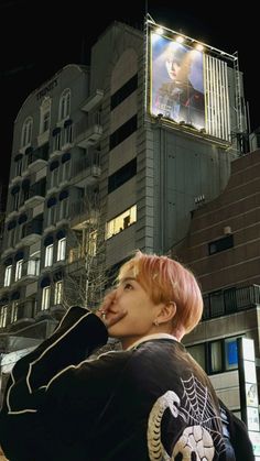 a person with pink hair standing in front of a building at night, wearing a black jacket