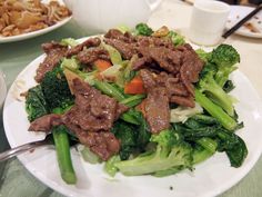 a white plate topped with beef and broccoli covered in sauce next to a bowl of noodles