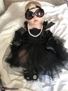 a baby wearing sunglasses and a black dress laying on a white sheet with a tiara