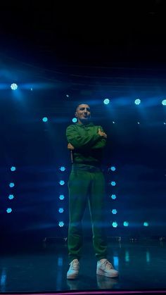 a man standing on stage with his arms crossed in front of the camera and lights behind him