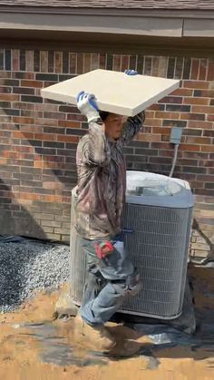 a man carrying an air conditioner on his head in the mud, with a brick wall behind him