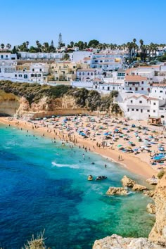 the beach is crowded with people and umbrellas in clear blue water, surrounded by white buildings