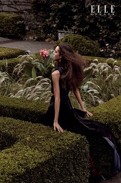 a woman sitting on top of a lush green hedge next to a pink flower in a garden