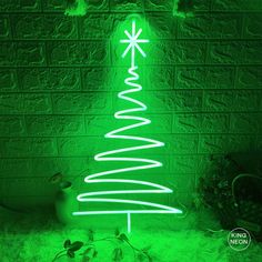 a green christmas tree is lit up in front of a brick wall with snow on the ground