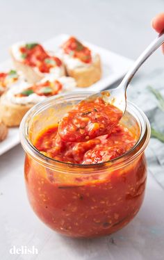 a spoon full of tomato sauce in a jar with bread on the table behind it