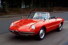 an old red sports car driving down the road with two people in the back seat