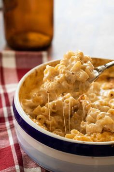 a spoon full of macaroni and cheese being lifted from a bowl with a red checkered table cloth