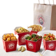 four take out boxes filled with food on top of a white table cloth next to chopsticks