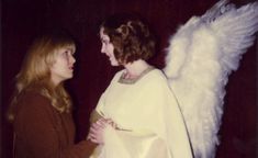 two women dressed in white with angel wings on their shoulders, facing each other and talking