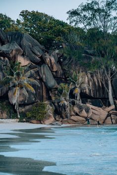 two people are standing on the beach near some rocks and palm trees in the background