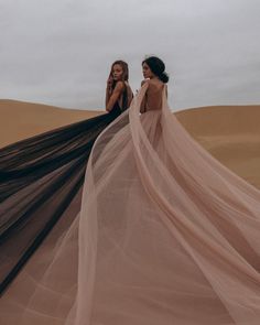 two women standing in the sand wearing long black and pink dresses with sheer fabric on them