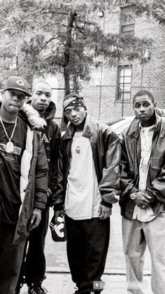 five young men are standing in front of a brick building, one is wearing a hat and the other wears a leather jacket