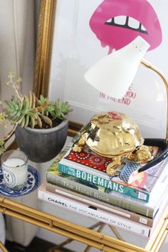 a table topped with books next to a potted plant and a gold framed mirror