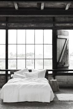 a bed with white sheets and pillows in front of large windows