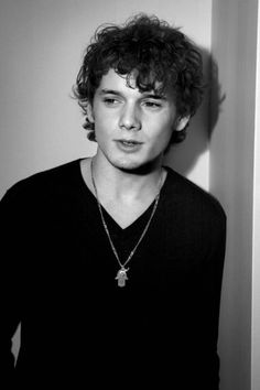 a young man with curly hair wearing a black shirt and silver necklace standing in front of a white wall