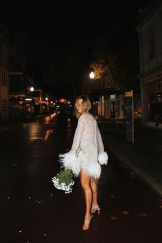 a woman walking down the street at night with flowers in her hand and feathers on her skirt