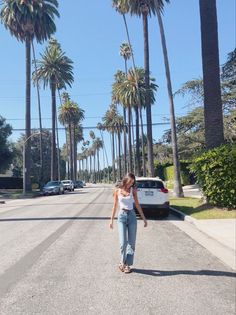 a woman is walking down the street in front of palm trees and a white car