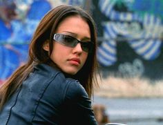 a woman wearing sunglasses sitting on a motorcycle in front of a graffiti covered wall and looking at the camera