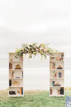 an open bookcase with flowers and books on it next to the beach in front of the ocean