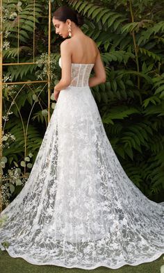 a woman in a white wedding dress standing next to some bushes and plants with her back turned towards the camera