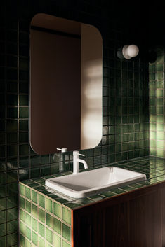 a white sink sitting under a mirror on top of a green tiled wall next to a wooden cabinet