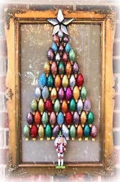 a christmas tree made out of colored eggs in a frame on a brick wall next to a small figurine