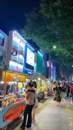 people are standing on the sidewalk in front of an outdoor food stand at night time